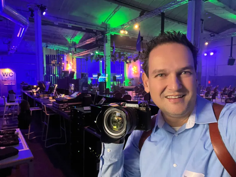 Fotograaf met camera maakt een selfie in een kleurrijk verlichte evenementenhal met een podium