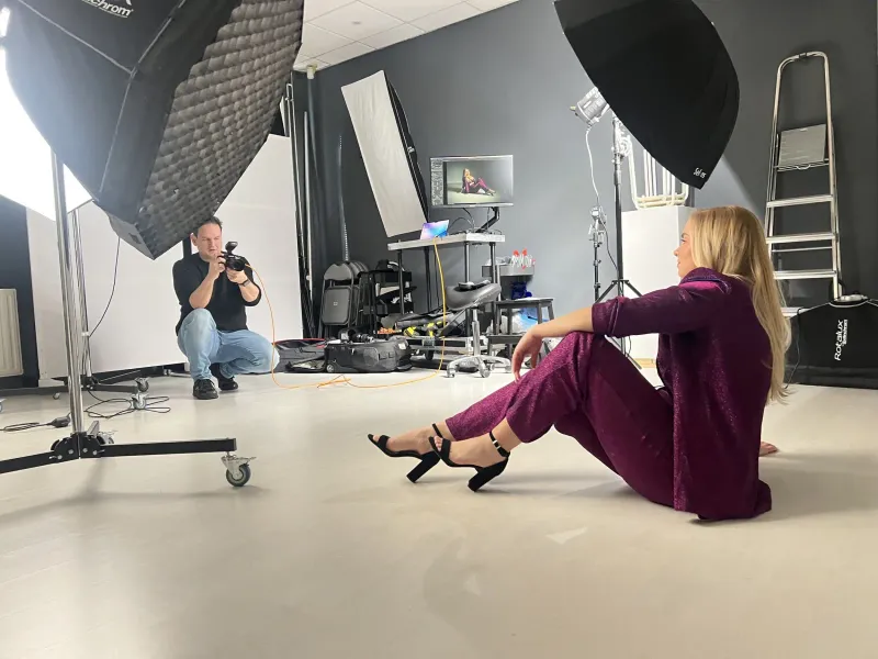 Fotograaf fotografeert in de studio een vrouw in een bordeauxrood pak die op de vloer zit, met softboxen eromheen