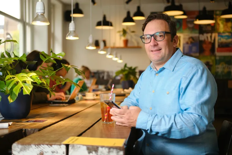 Lachende man met een glas thee aan een lange houten tafel in een kantoor vol planten