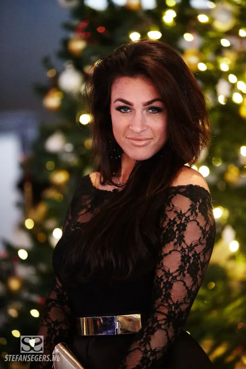 Vrouw in een zwarte kanten top poseert voor een versierde kerstboom tijdens een evenement
