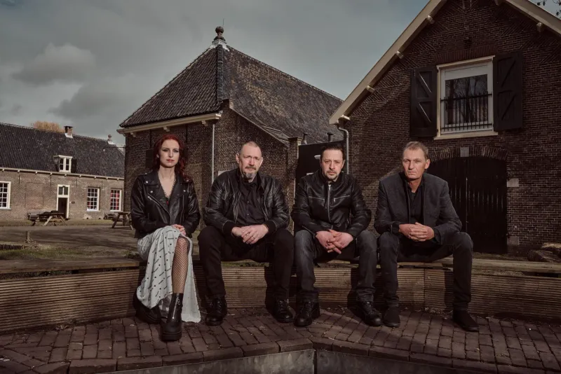 Bandportret van vier mensen in leren jassen op een muurtje voor een donkere loods