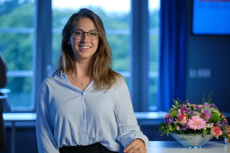 Lachende vrouw met bril in een lichtblauwe blouse aan een tafel met een boeket voor grote ramen
