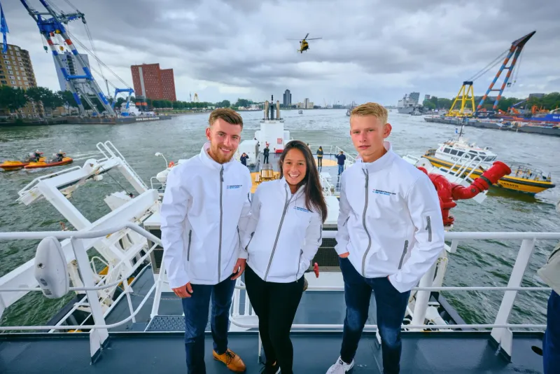 Drie bemanningsleden in witte crewjassen op een scheepsdek met havenkranen en een reddingshelikopter