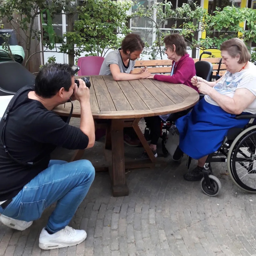Een man neemt een foto van twee vrouwen die op een houten tafel zitten, een vrouw met een rolstoel en een oudere vrouw die hand in hand zitten en een gesprek voeren.
