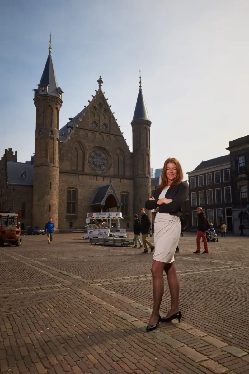 Vrouw in zakelijke kleding op een historisch plein met torens en een gotische zaal op de achtergrond