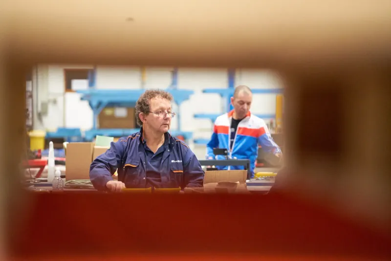 Man in blauwe overall werkt aan een werkbank in een werkplaats, met een collega op de achtergrond