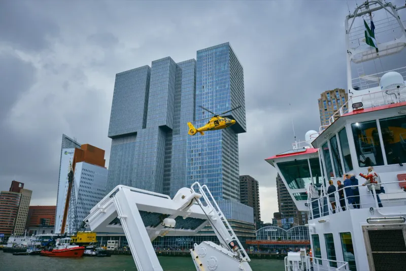 Gele reddingshelikopter scheert langs de hoekige wolkenkrabbers van De Rotterdam, gezien vanaf een schip