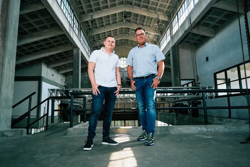 Twee mannen op een stalen loopbrug in een grote lege industriehal met hoge ramen