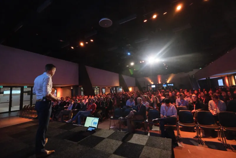 Spreker op een podium voor een zittend publiek in een verduisterde zaal met tegenlicht