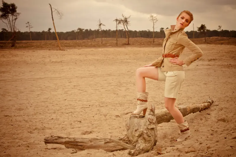Modeportret van een vrouw die met een voet op een boomstam poseert op een zandvlakte met kale bomen