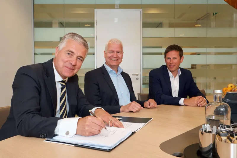 Drie mannen in pak aan een vergadertafel terwijl een van hen een document ondertekent
