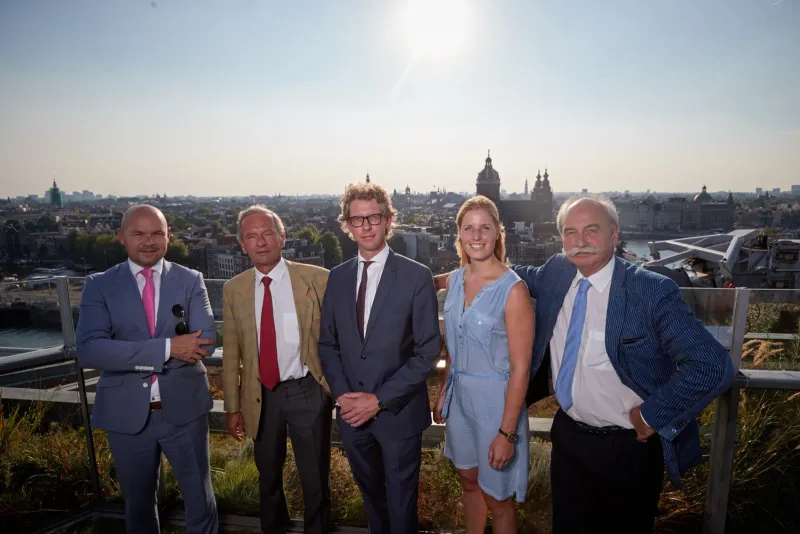 Vijf mensen in zakelijke kleding op een dakterras met de skyline van Amsterdam bij zonsondergang