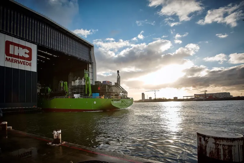 Groen vrachtschip voor een grote scheepswerfhal onder een dramatische lucht boven het water