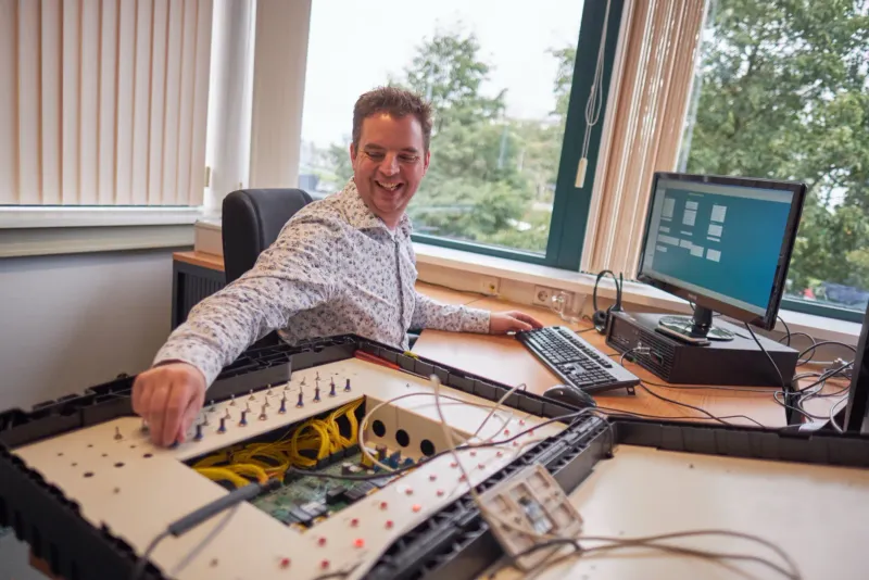 Man aan een bureau met monitoren en een geopende glasvezelkast op de voorgrond