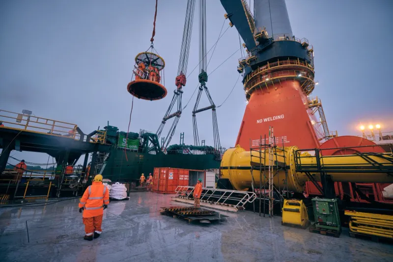 Kraan hijst een grote gele kabelhaspel aan boord van een offshoreschip terwijl bemanning in oranje werkkleding meekijkt
