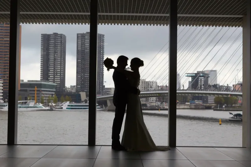 Silhouet van een bruidspaar voor een groot raam met uitzicht op de skyline en de Erasmusbrug