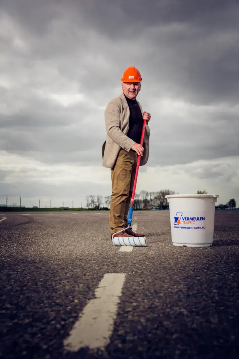 Man in colbert met bouwhelm, verfroller en emmer op een lege weg met windmolens op de achtergrond