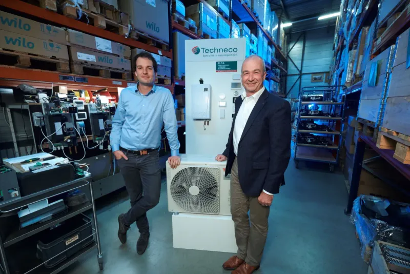 Twee mannen naast een warmtepomp in een magazijngang met blauwe stellingen
