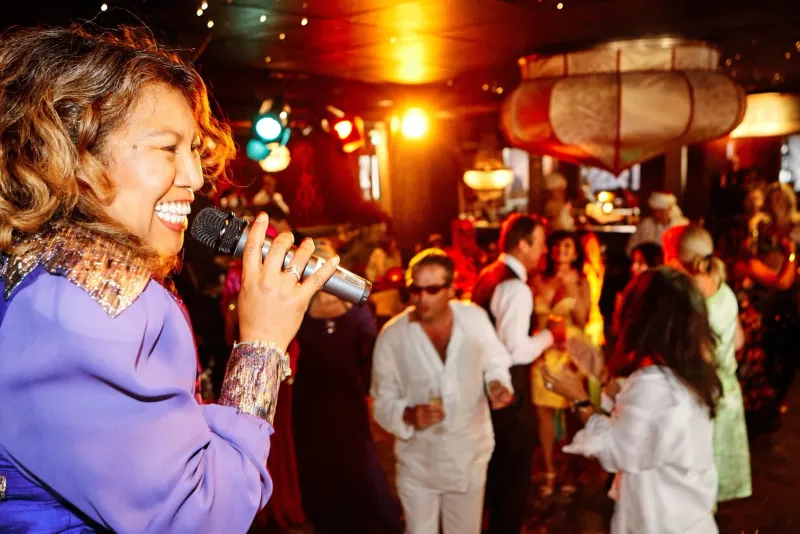 Vrouw in het paars zingt in een microfoon op een sfeervol verlicht feest met dansende gasten