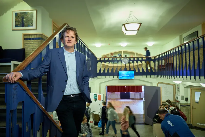 Man in colbert leunt op een trapleuning in een schoolgebouw met lockers