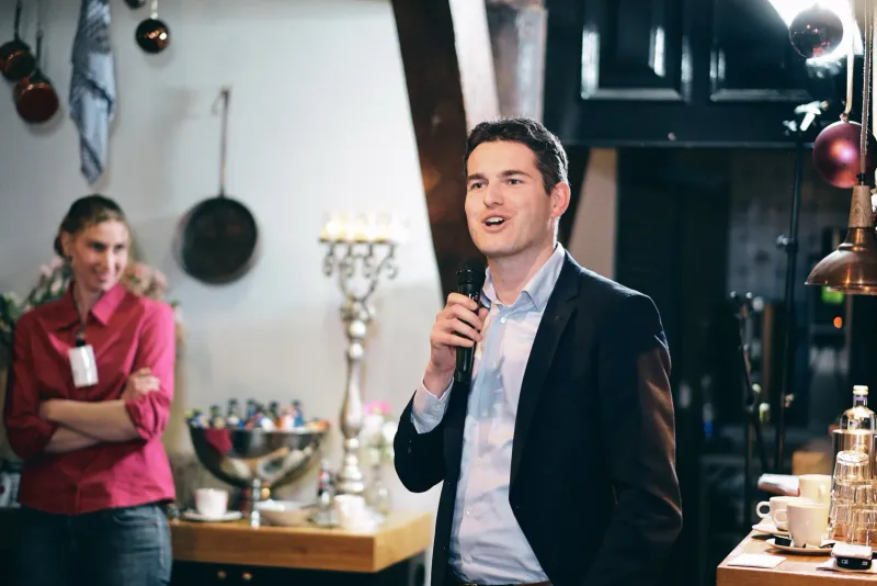 Man met microfoon spreekt in een sfeervolle keuken met koperen pannen terwijl een vrouw toekijkt