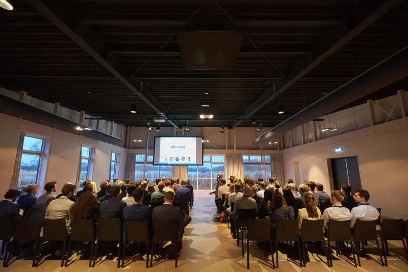 Congreszaal met rijen toehoorders gericht op een verlicht podium en scherm