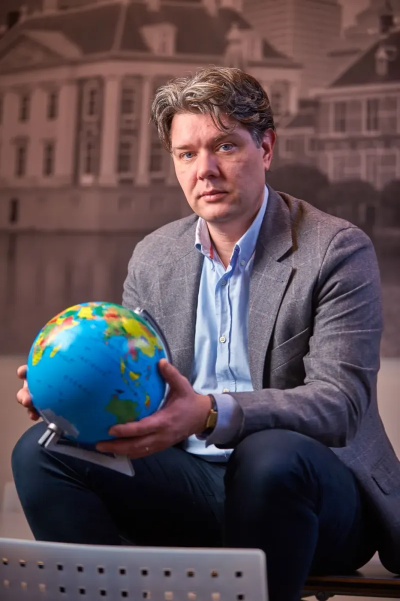 Man in grijs colbert hurkt met een kleine wereldbol voor een geprinte achtergrond van een monumentaal gebouw