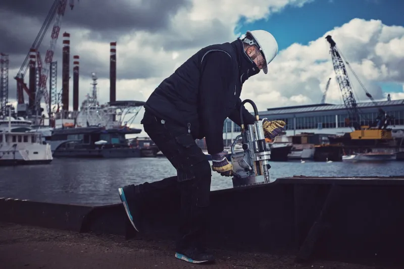 Werknemer met helm bedient apparatuur op een ponton met havenkranen en schepen op de achtergrond