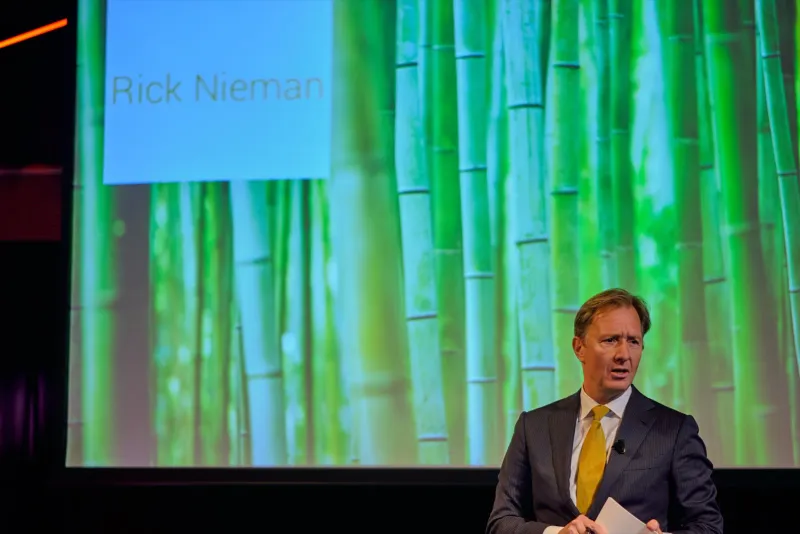 Dagvoorzitter spreekt achter een katheder voor een groene achtergrond met bamboemotief