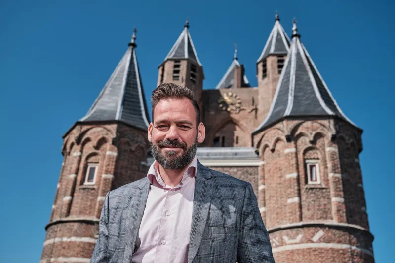 Lachende man in geruit colbert voor de twee bakstenen torens van een neogotische kerk