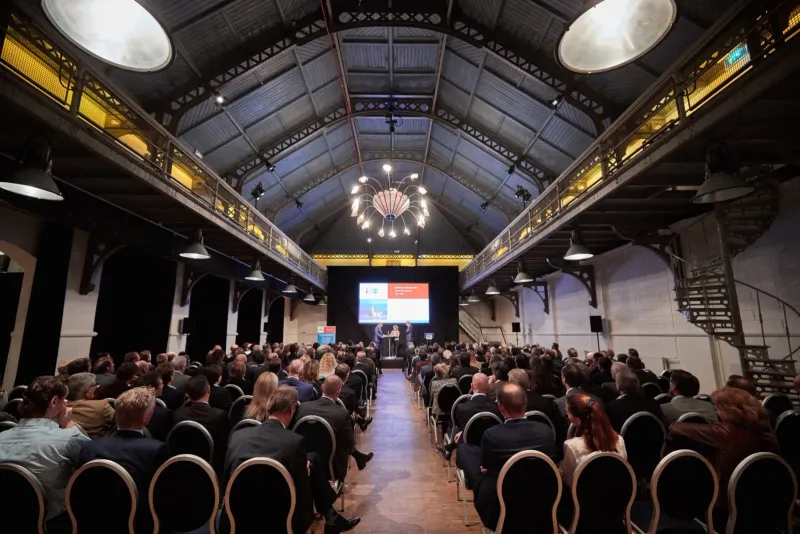 Congreszaal in een industrieel monument met stalen dakspanten en publiek dat naar een verlicht podium kijkt