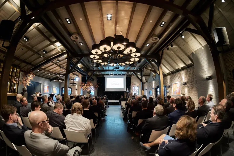 Publiek in een tongewelfde zaal kijkt naar een presentatie onder een kroonluchter