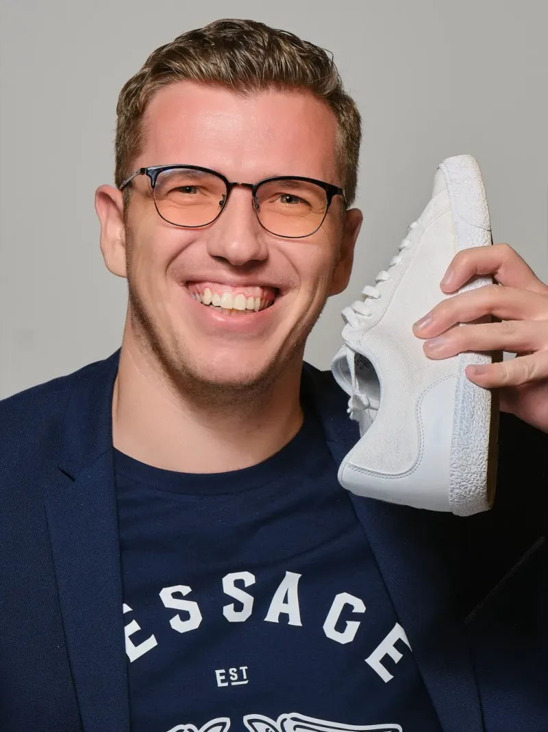Lachende jonge man in colbert houdt een witte sneaker naast zijn gezicht