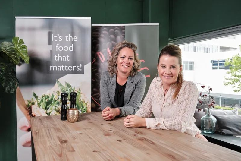Twee vrouwen aan een houten tafel naast een banner met de tekst It's the food that matters